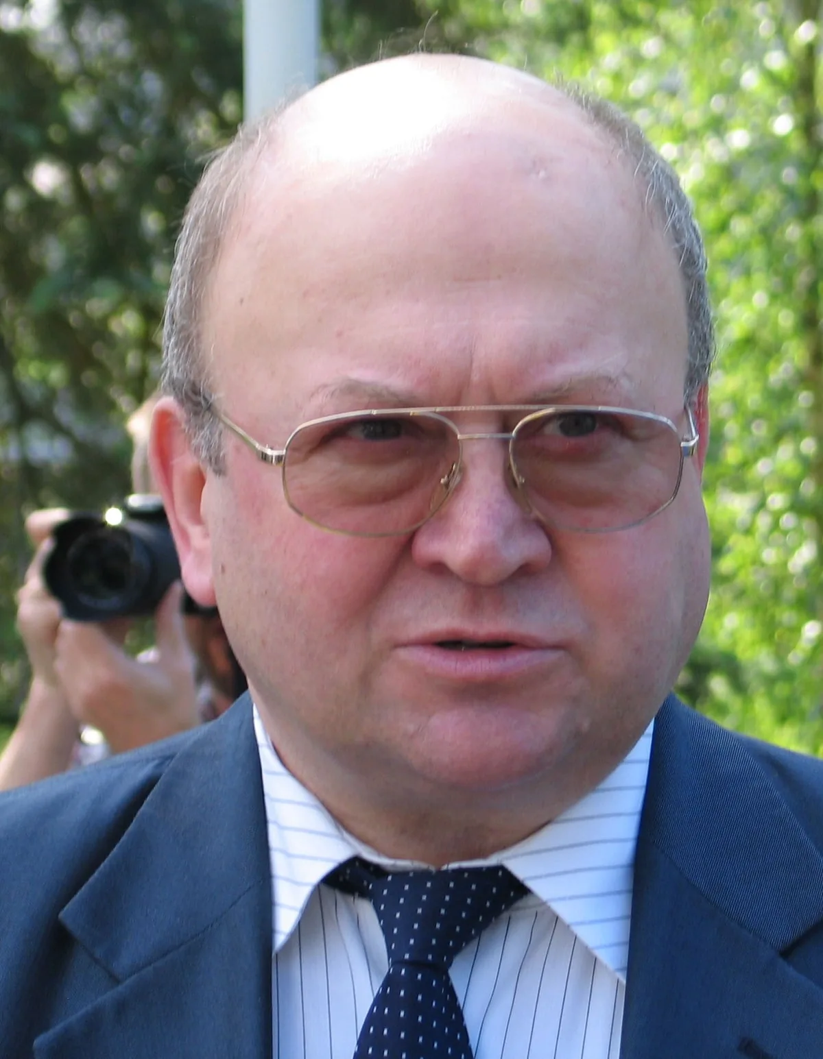 Vladimír Remek wearing glasses, a dark jacket and tie in an outdoor portrait