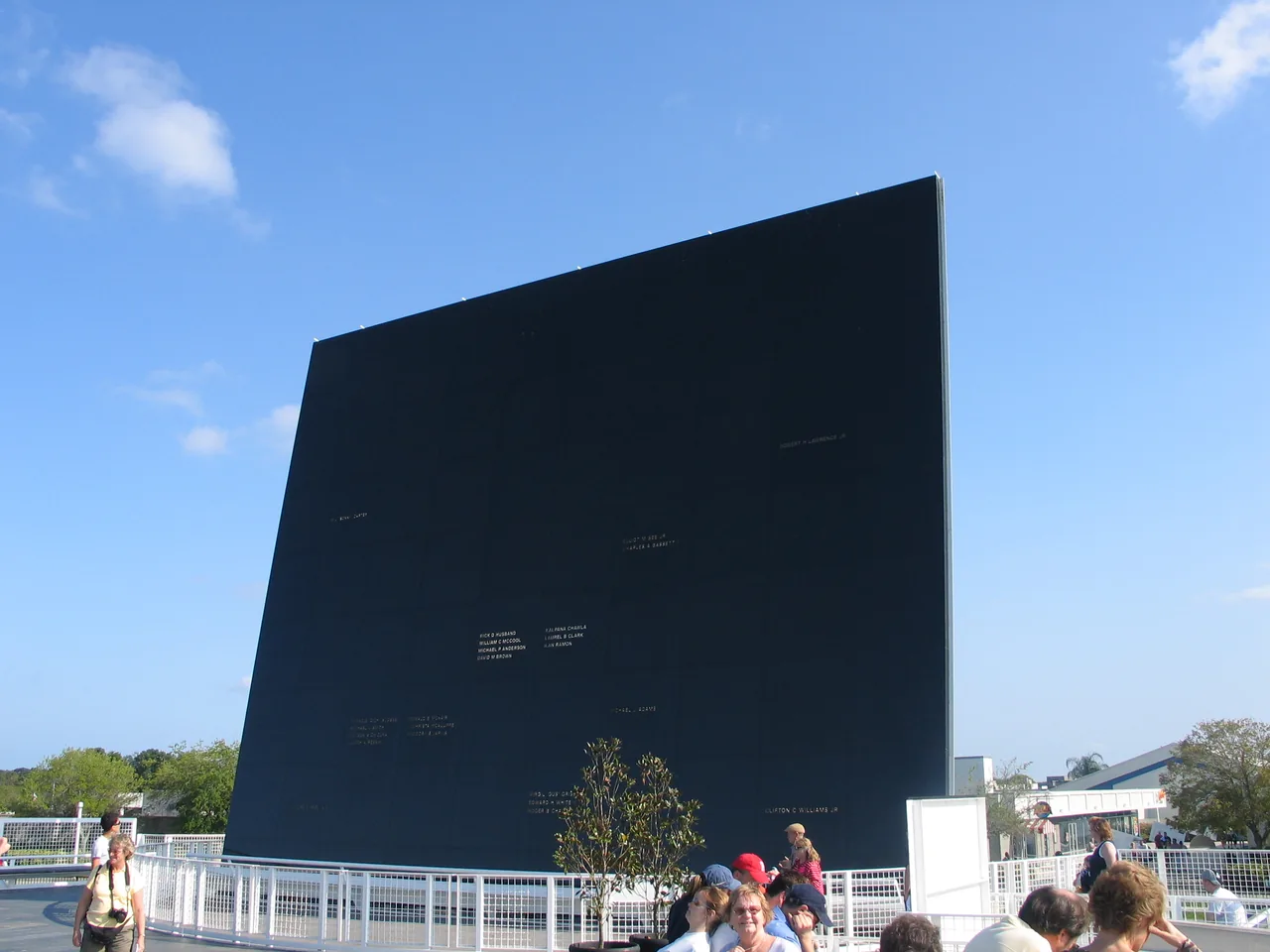 A large dark memorial panel rises above visitors beneath a blue sky
