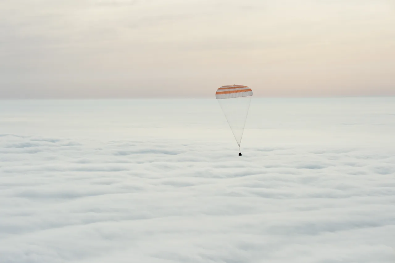 An orange-and-white parachute suspends a small dark Soyuz capsule above a broad white cloud layer.