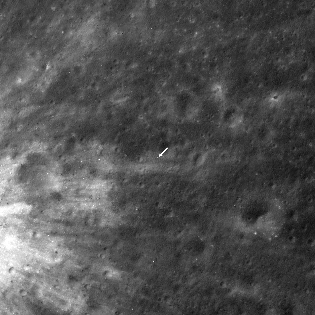 A grayscale orbital view of cratered lunar ground, with a white arrow identifying the tiny SLIM lander near the center.