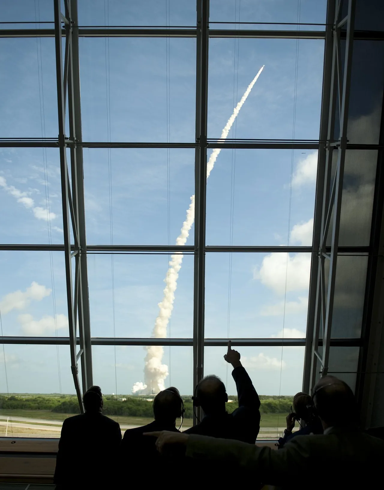 People watch a distant rocket and long white exhaust plume through a large control-room window