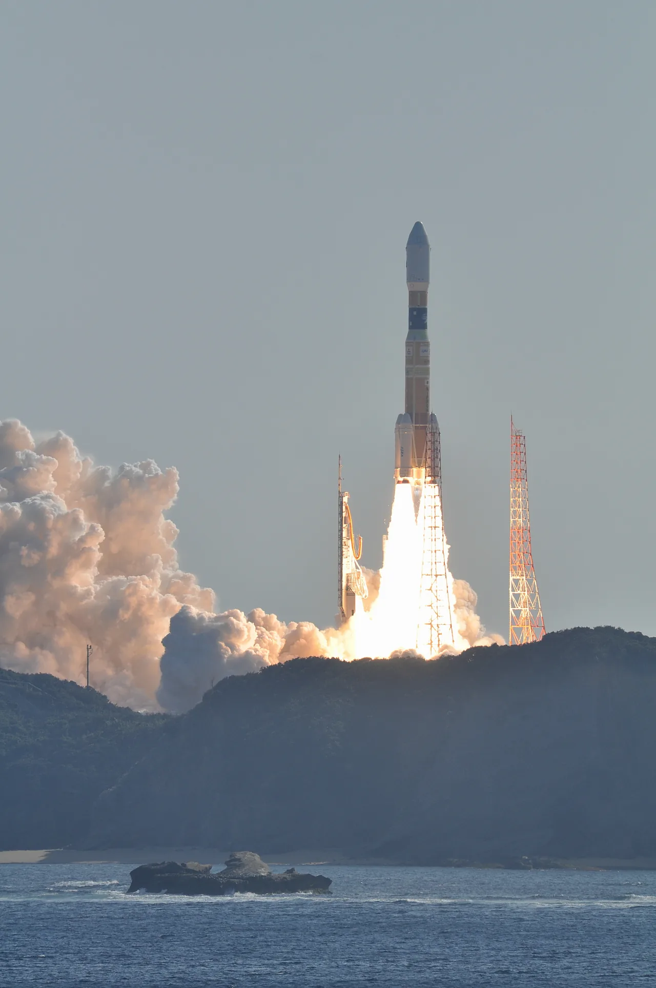 H-IIB climbs above Tanegashima in daylight, with flame, launch towers, wooded hills and sea visible in the full portrait frame.
