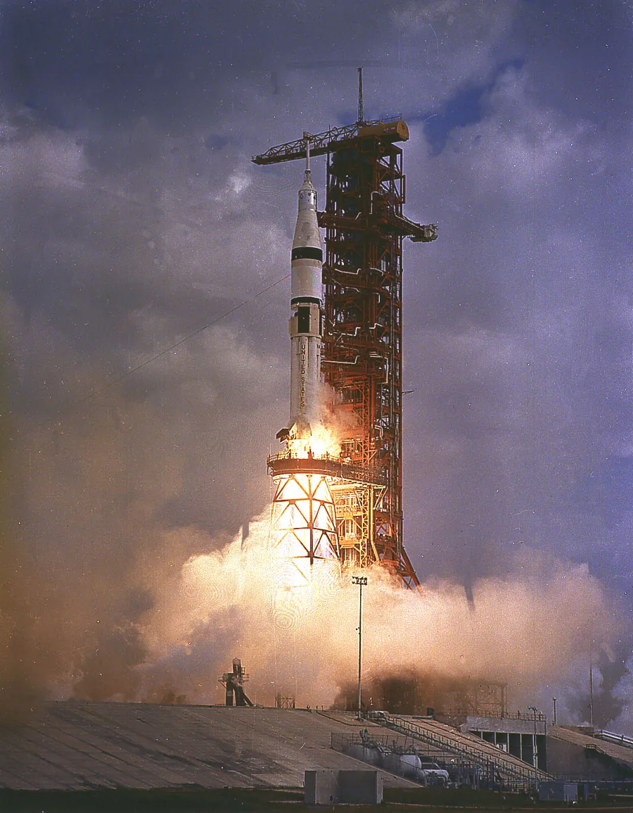 A Saturn IB rises beside the launch tower through bright exhaust and smoke