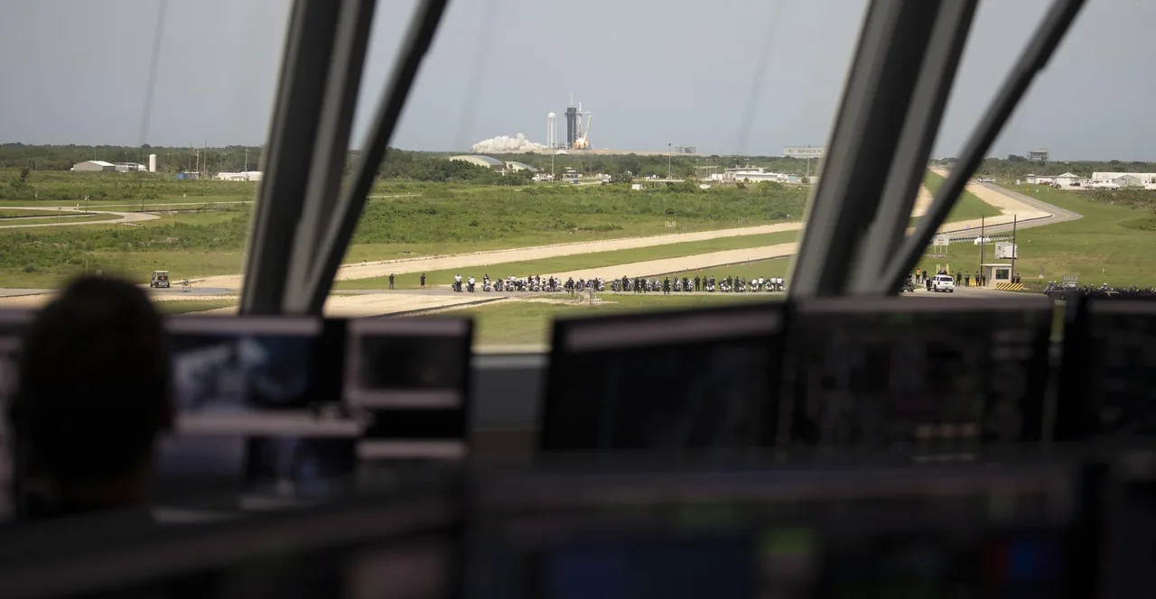 Through launch-control windows and above foreground monitors, Falcon 9 lifts off in the distance with Crew Dragon.