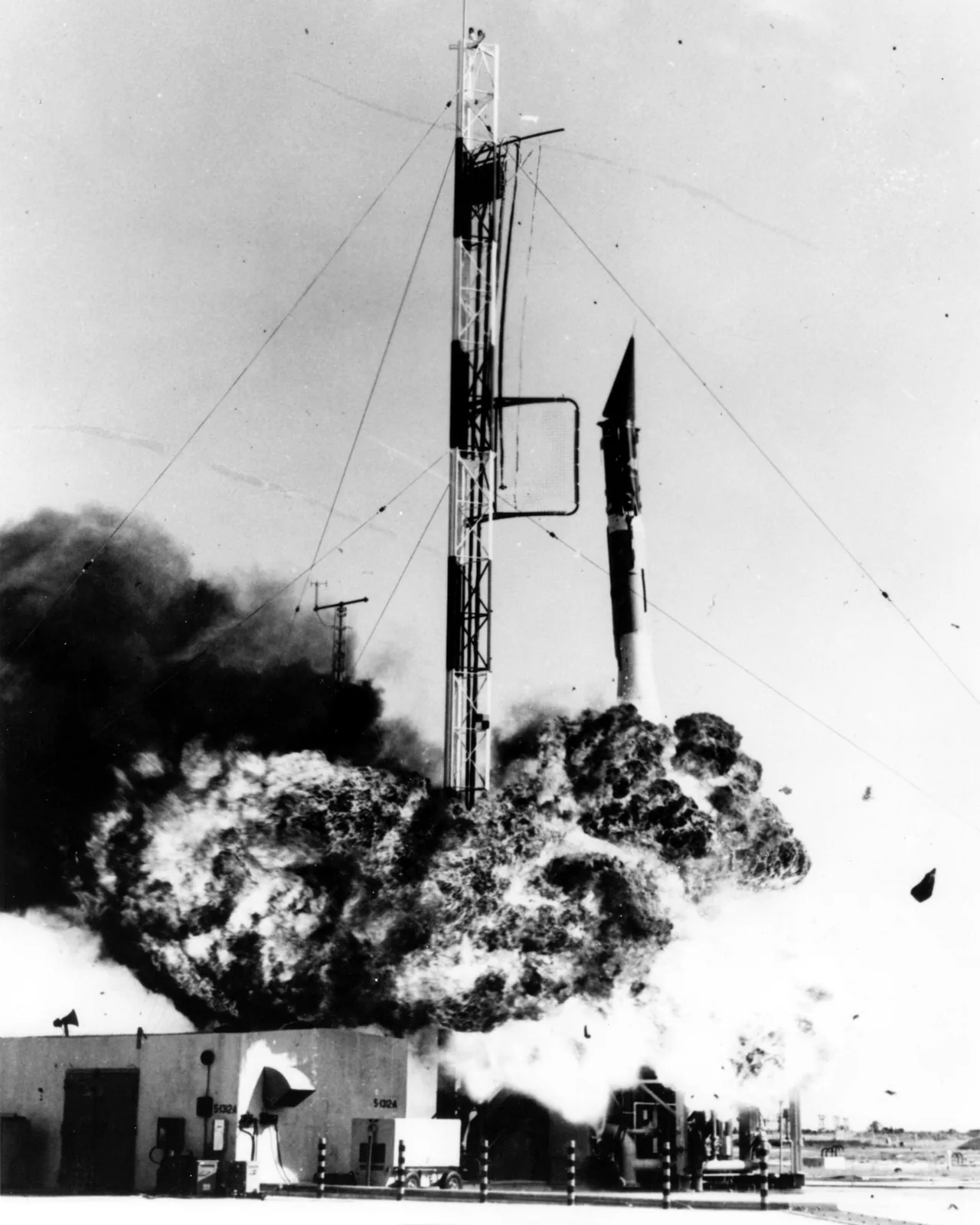Flames and smoke surround the base of the Vanguard TV-3 rocket beside its launch tower