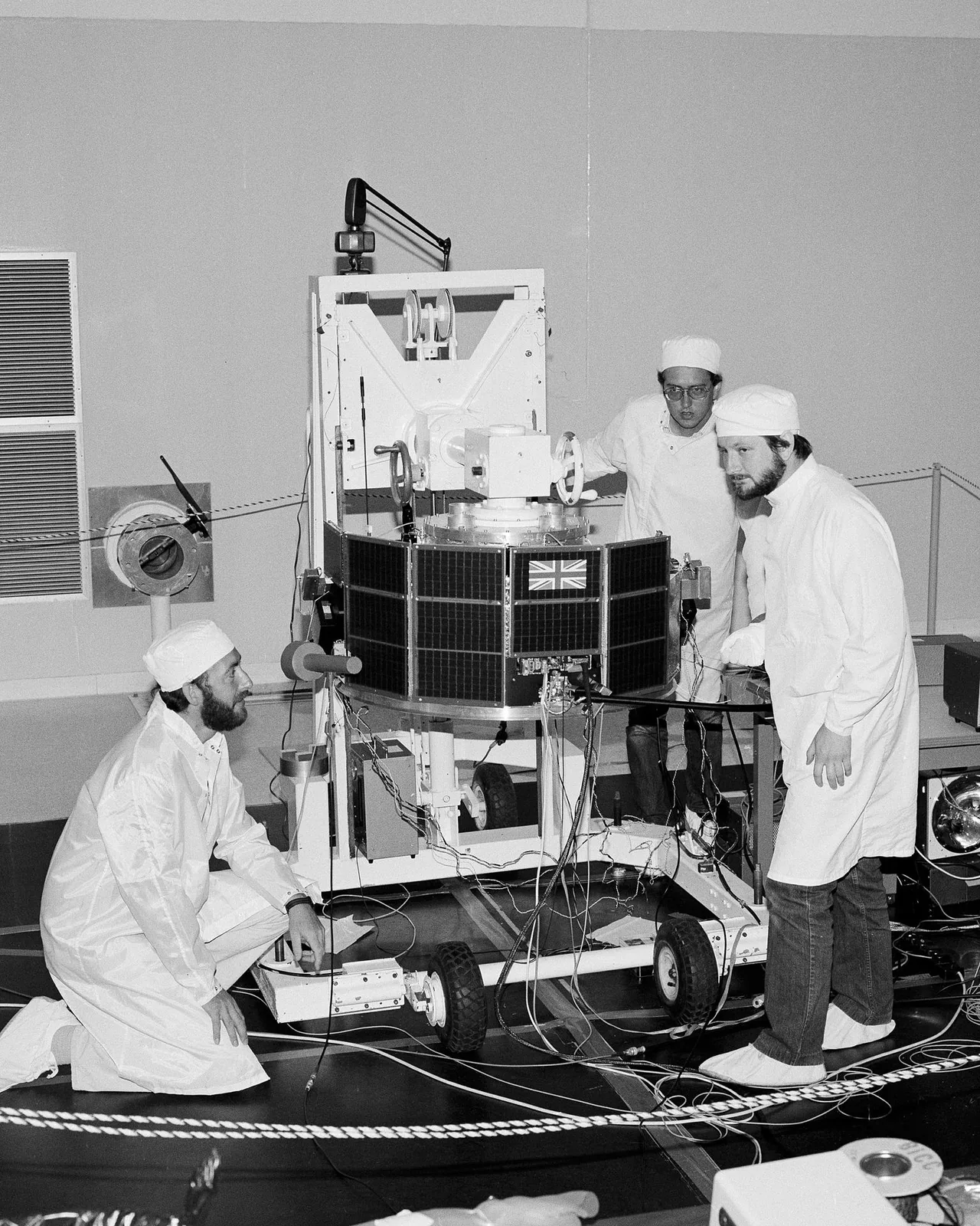 Three technicians work around the AMPTE UK spacecraft, whose cylindrical body bears a British flag