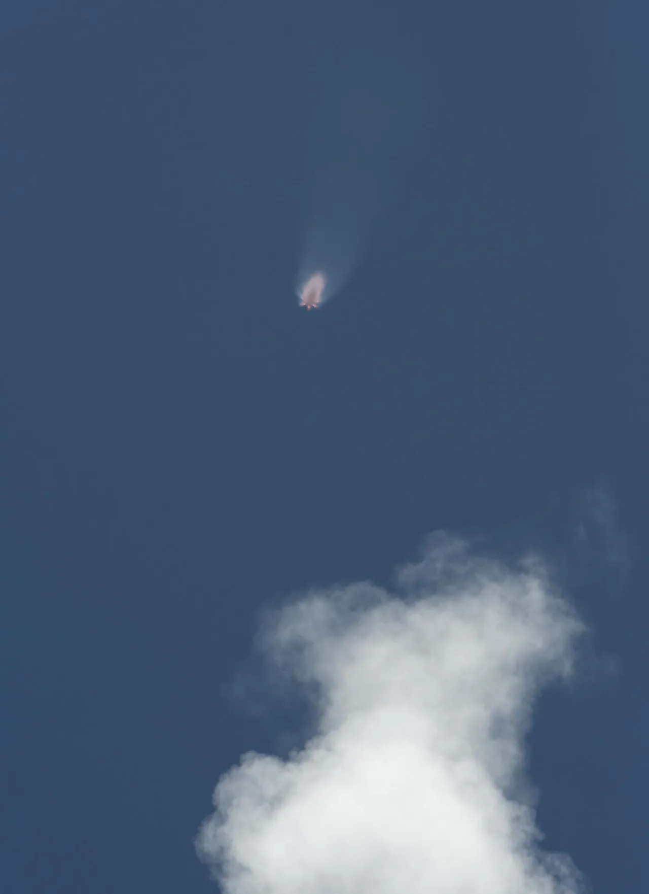 A distant Falcon 9 climbs through blue sky above a bank of cloud
