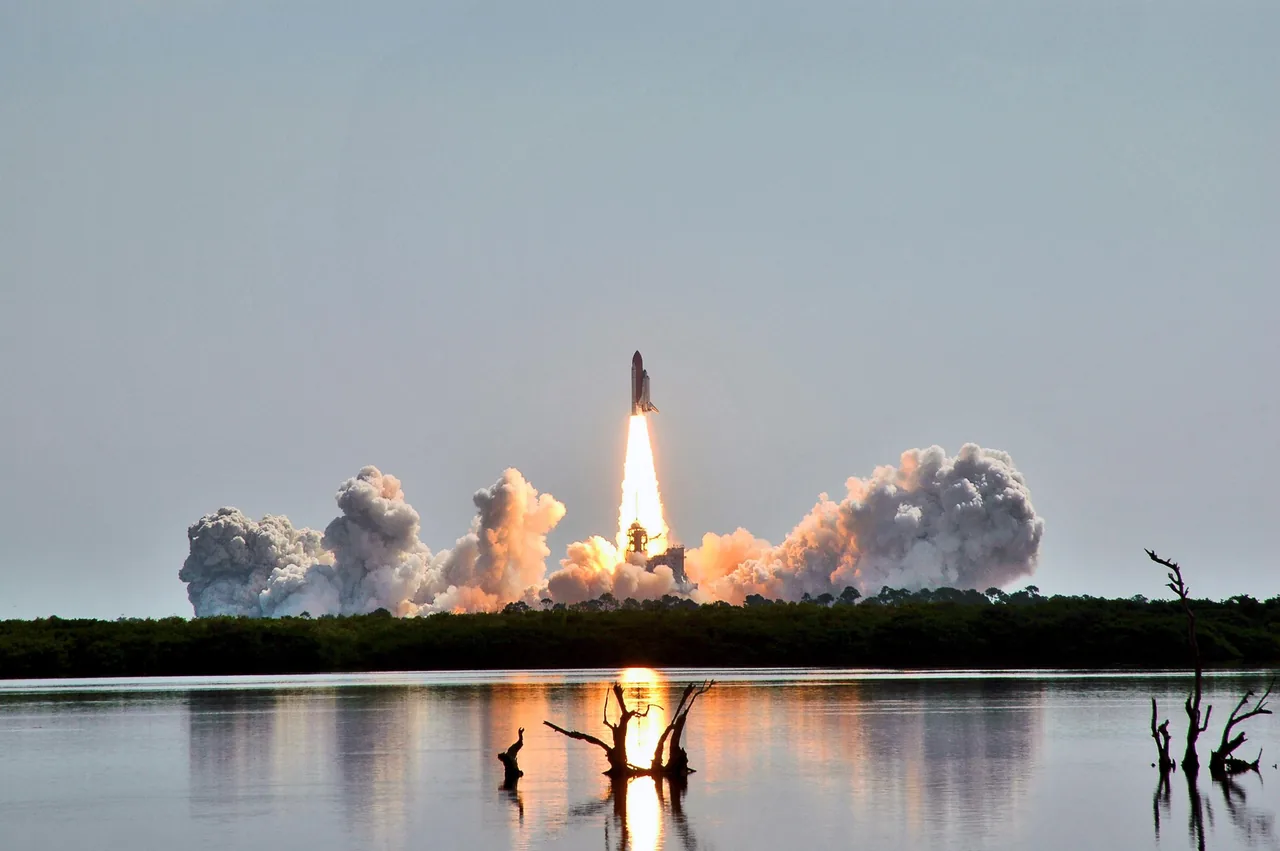 Discovery rises above its launch pad on a bright exhaust plume, with billowing clouds, trees, and a water reflection below.