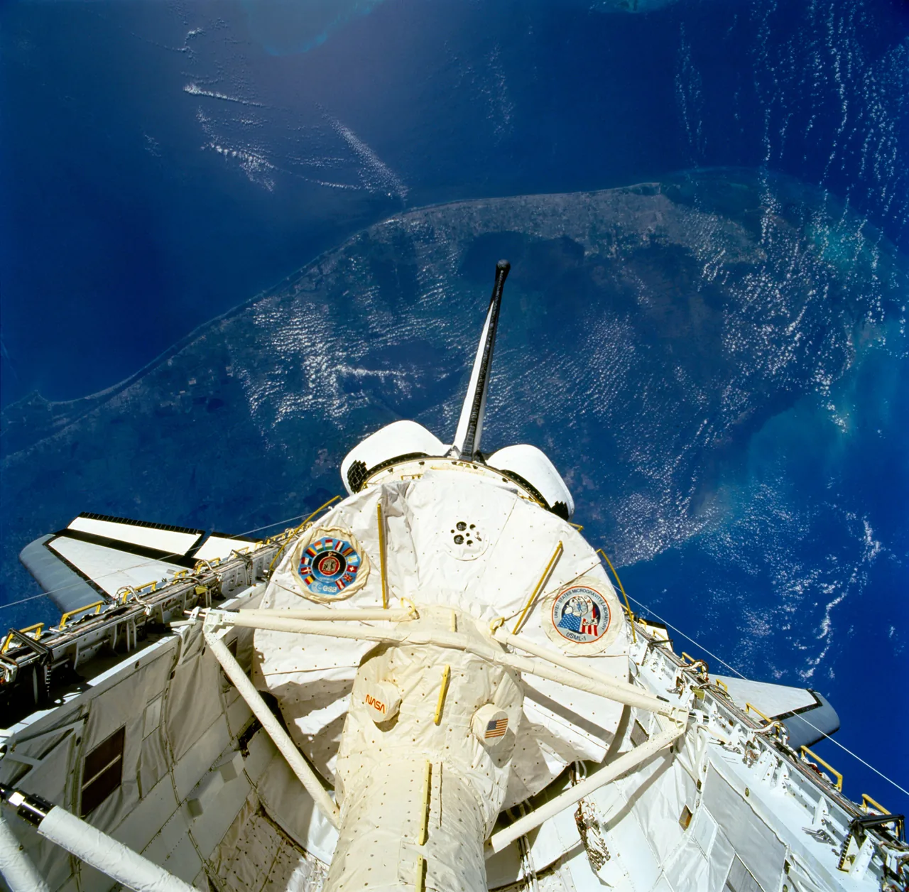 The white USML-1 laboratory module fills Columbia’s open payload bay, with blue ocean and coastal land below