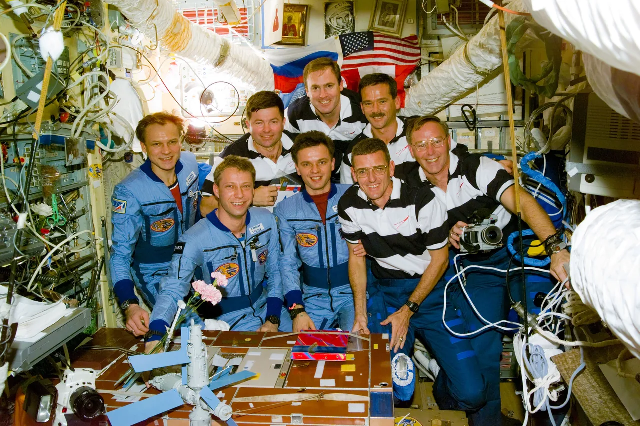 Eight STS-74 and Mir-20 crew members pose together inside Mir, surrounded by equipment and flags.