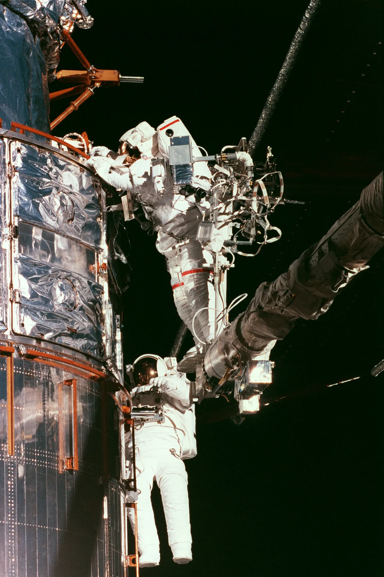 Two astronauts in white suits work beside Hubble’s curved silver exterior against black space, with the upper astronaut supported by the shuttle’s robotic arm.