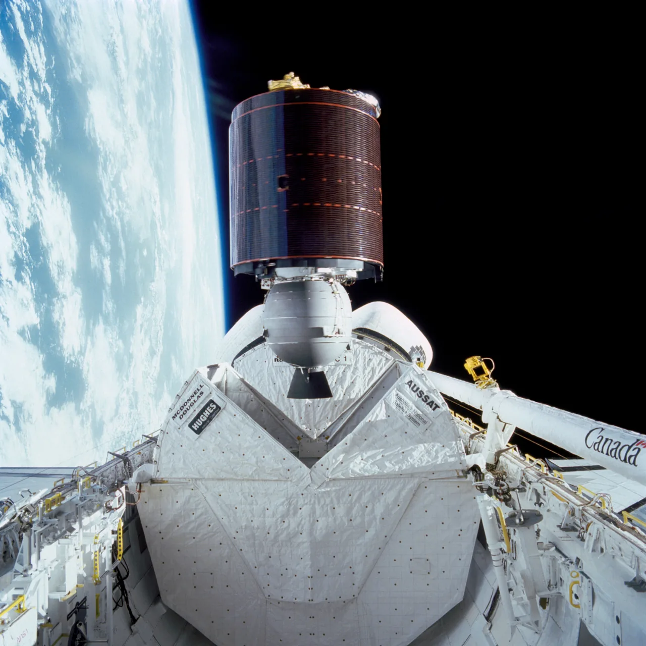 Cylindrical AUSSAT satellite with its attached propulsion module above Discovery’s open payload bay, with Earth beyond