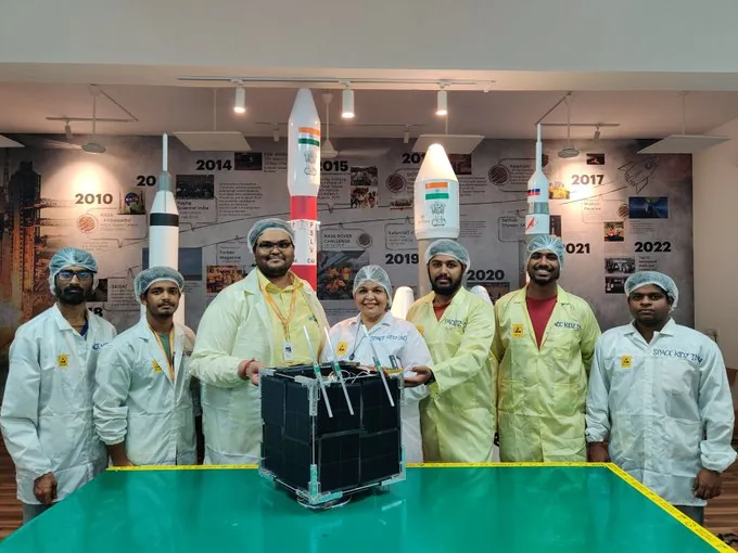Seven SpaceKidz India team members in cleanroom clothing stand behind the black box-shaped AzaadiSAT-2 spacecraft on a green work surface, with program displays and rocket models behind them.