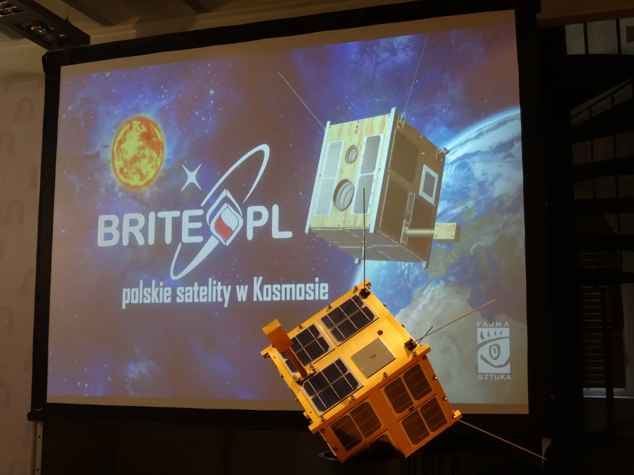 A gold Heweliusz satellite model displayed in front of a BRITE-PL presentation screen at an indoor event