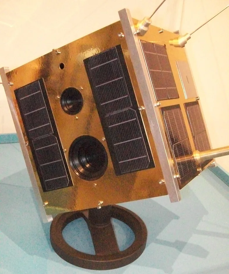Gold-colored Lem satellite model on a round display stand, with black solar cells and two visible optical openings