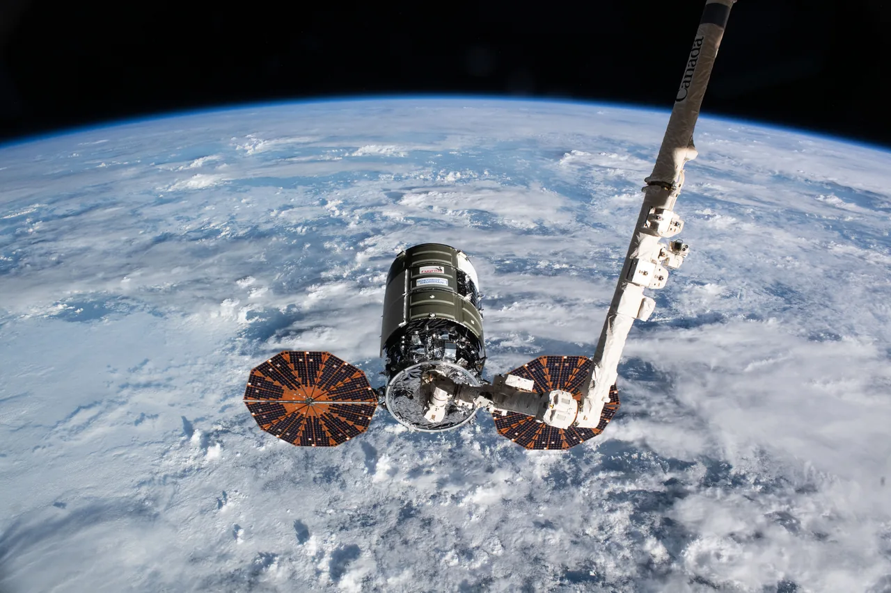 The cylindrical Cygnus cargo spacecraft and its two deployed circular solar arrays hang from Canadarm2 above Earth's curved, cloud-covered horizon.