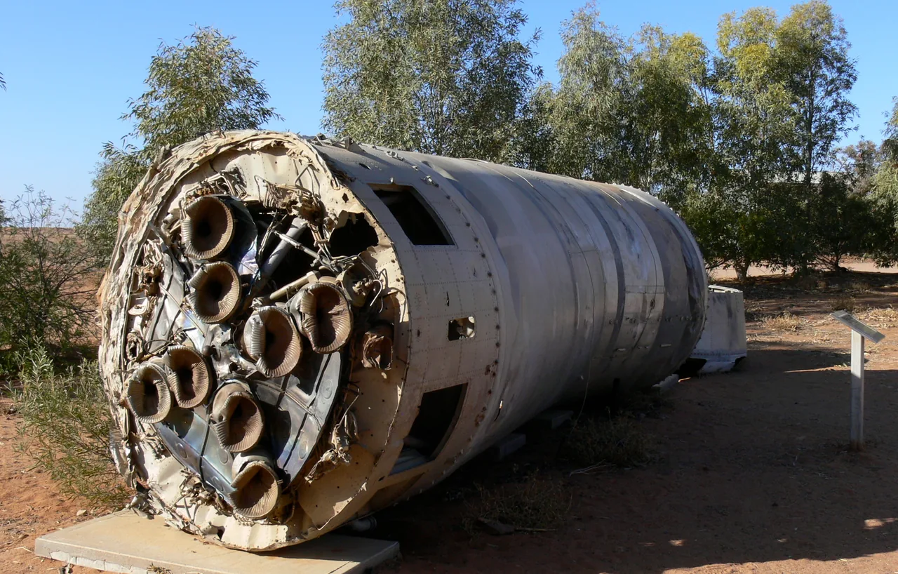A recovered cylindrical rocket stage lies outdoors with eight engine nozzles visible at its weathered end