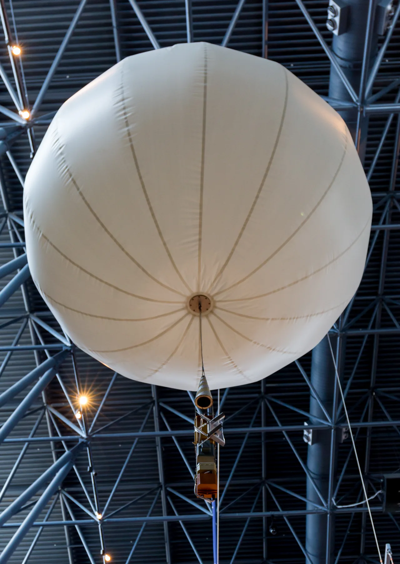 A pale round balloon with seams and a small suspended gondola hangs below a museum roof