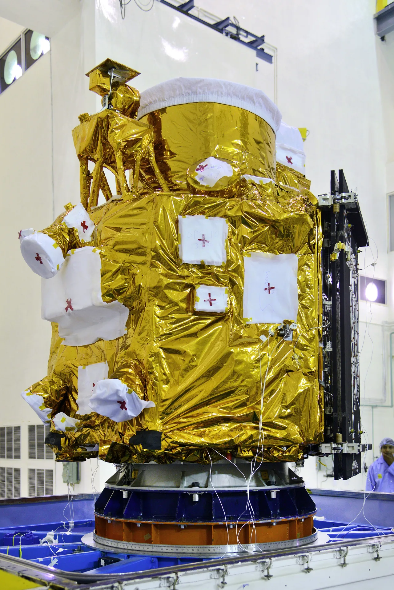 A gold-insulated satellite with protected fittings and a folded dark solar panel stands in a white cleanroom