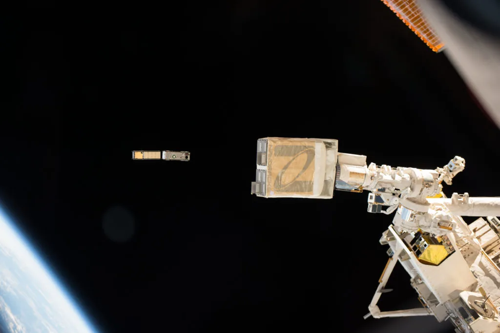 Small deployed objects near a station-mounted deployer, with Earth visible below