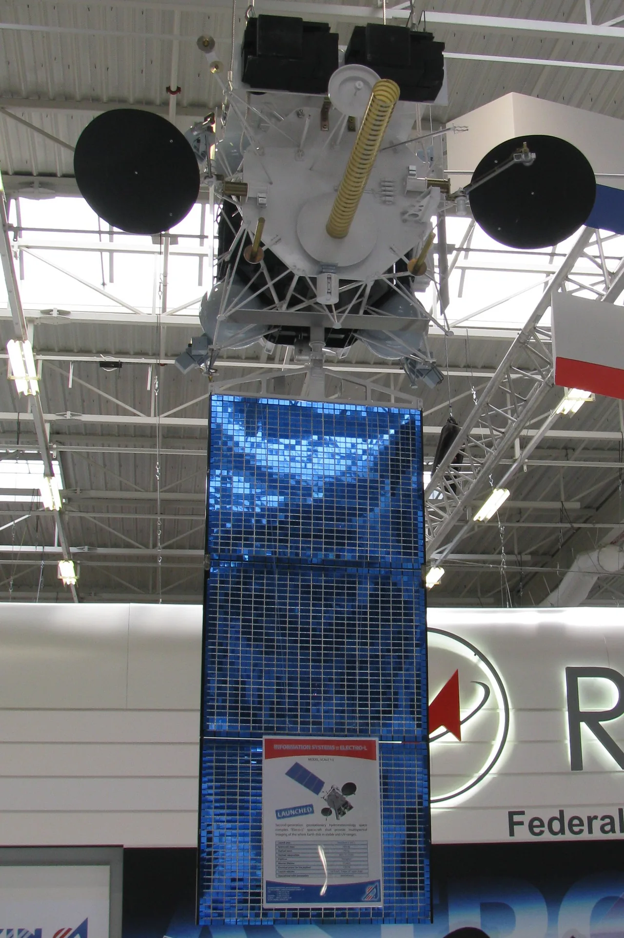 An Elektro-L model suspended above an exhibition display with two black dishes and a long blue solar panel