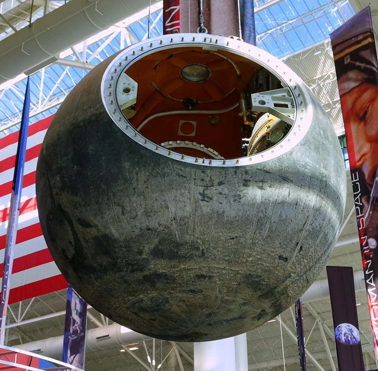 A dark spherical Foton return capsule hangs in a museum, with its open hatch and interior fittings visible