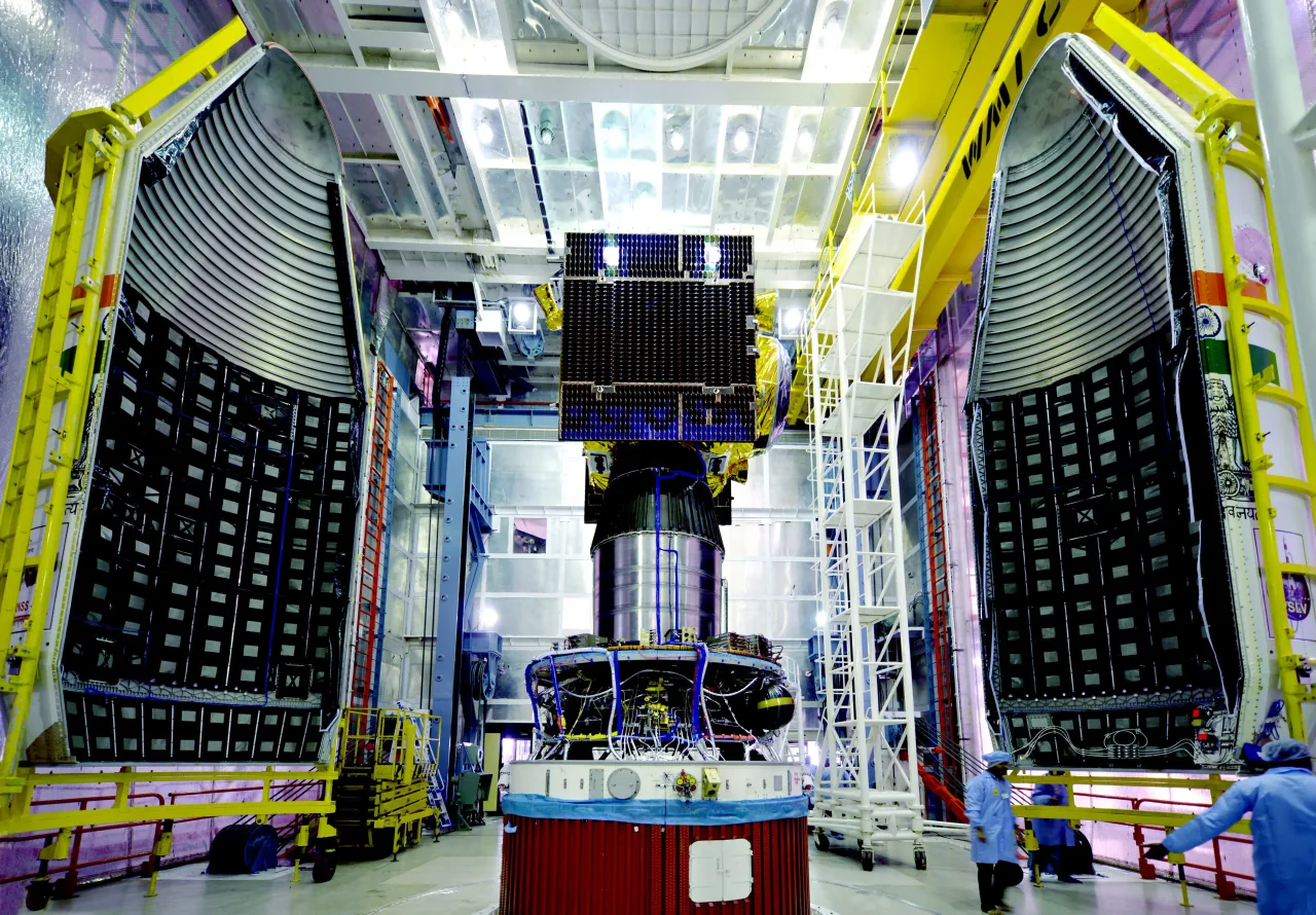 A gold-covered navigation satellite with a dark solar panel stands above its launch stage between open service-tower doors