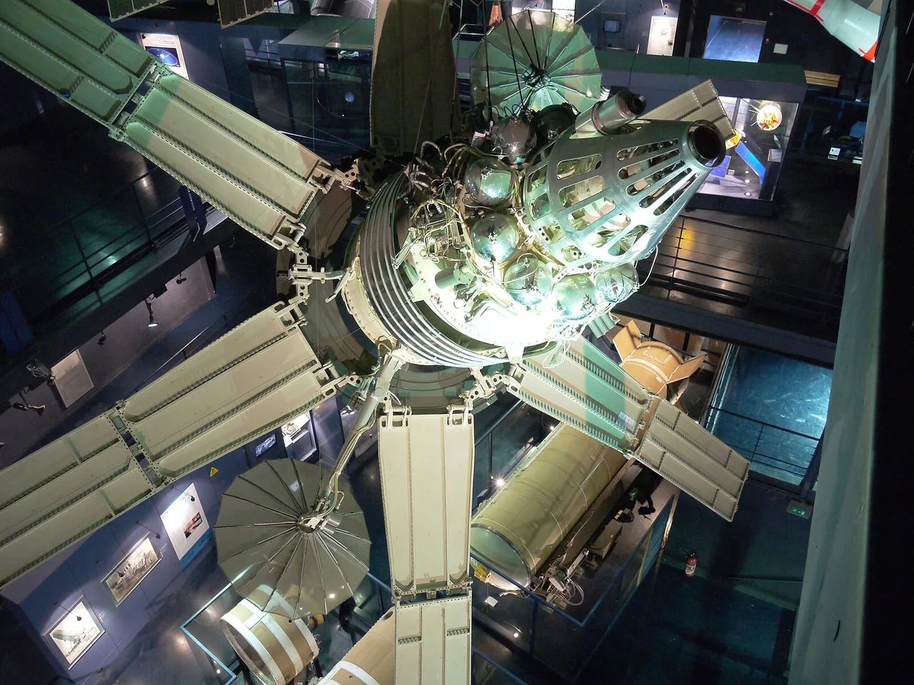 Full-scale Molniya 1A satellite model in a museum, with six pale solar-panel wings and dish antennas surrounding its central body.