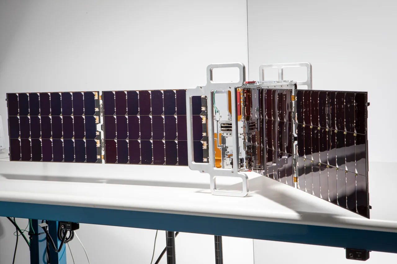 A PREFIRE CubeSat sits on a laboratory table with long dark solar arrays extending from its central structure