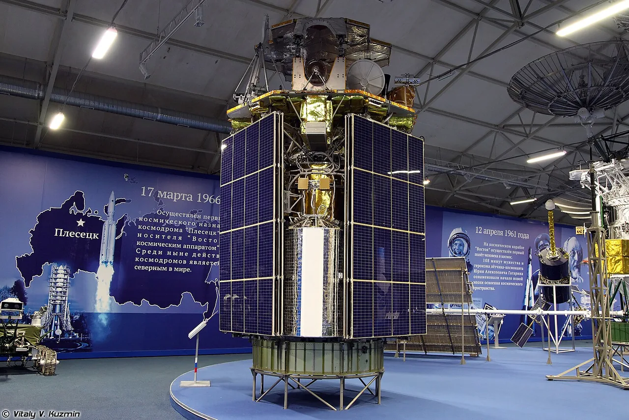 Tall cylindrical Raduga-1M display spacecraft indoors, wrapped in gold insulation with blue solar panels and multiple dish antennas.