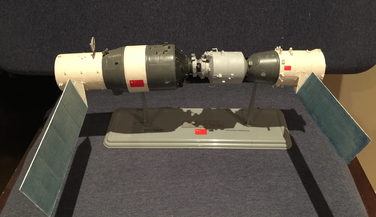 Photograph of a tabletop physical model showing a cylindrical Chinese Tiangong space laboratory with two solar panels docked nose-to-nose with a Shenzhou spacecraft model.