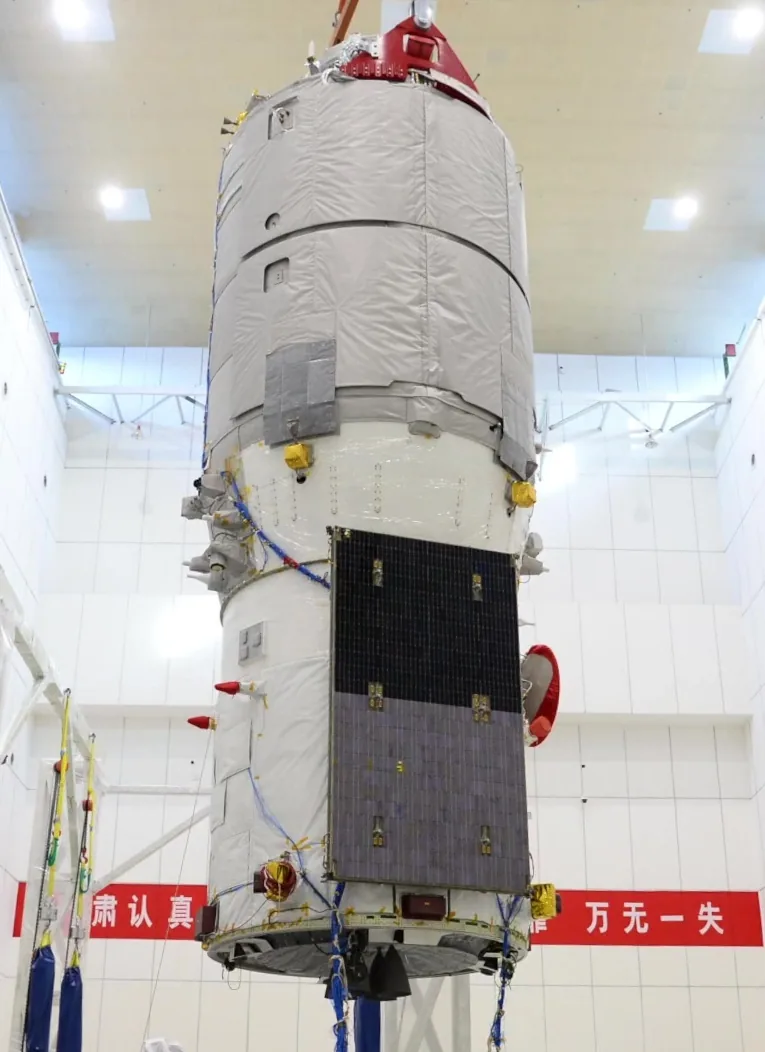 The cylindrical Tianzhou-2 cargo spacecraft standing vertically in a bright clean room, with insulation blankets, solar cells, wiring, and docking hardware visible.