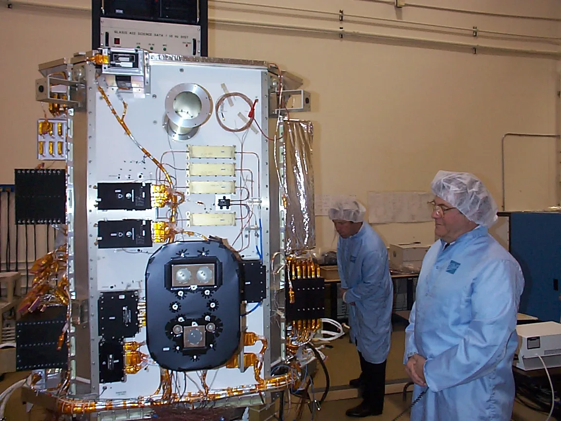 Technicians in blue garments stand beside the original ICESat spacecraft during integration