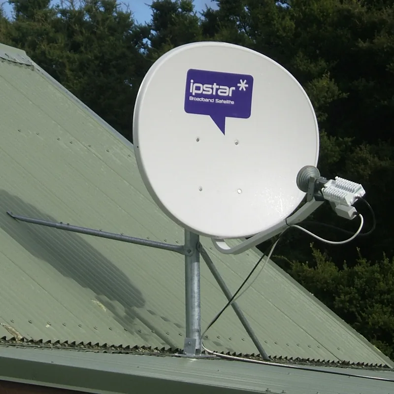 A white IPSTAR satellite dish stands beside a green corrugated roof with trees behind it