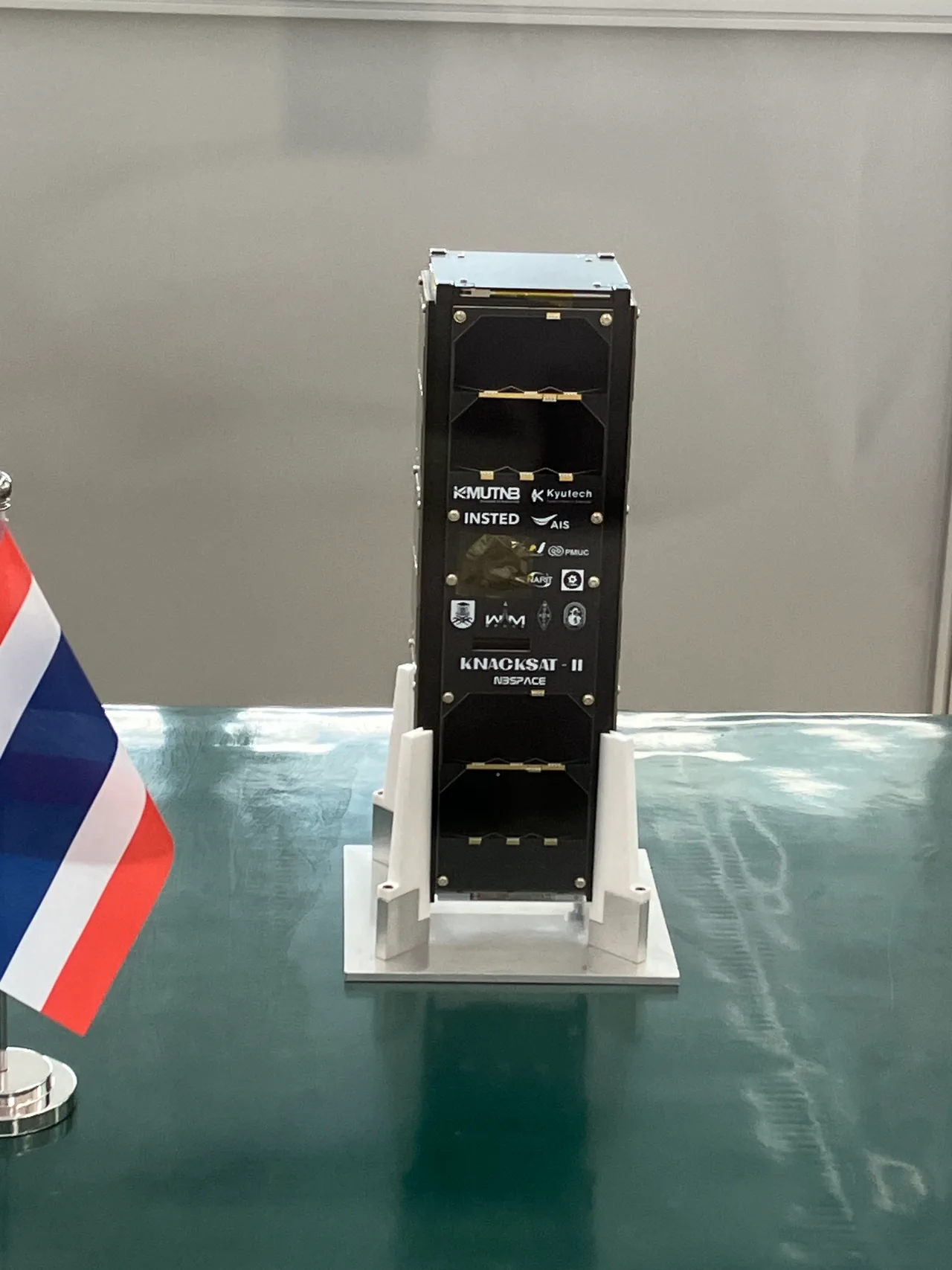 A tall black KNACKSAT-2 CubeSat stands on a white support beside a Thai flag in a clean room