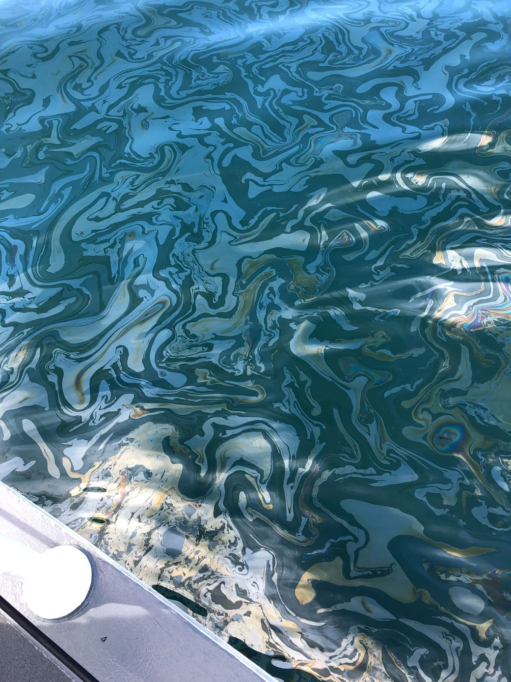 Swirling blue, silver and rainbow-colored oil sheen on seawater beside the edge of a small boat.