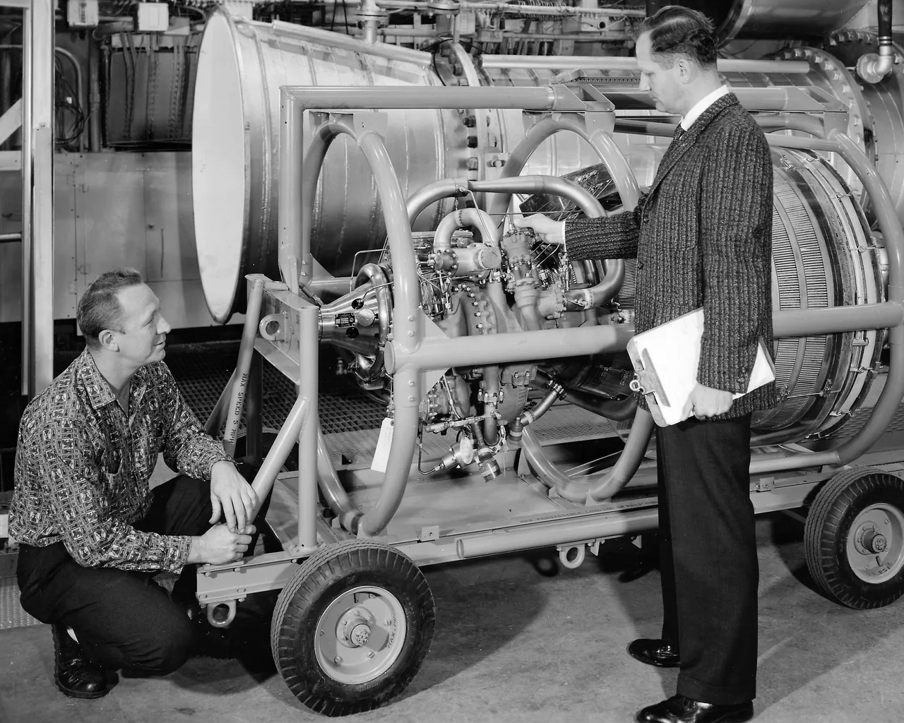 Two researchers examine an early RL10 engine on a wheeled laboratory support, with pipes and a large nozzle visible