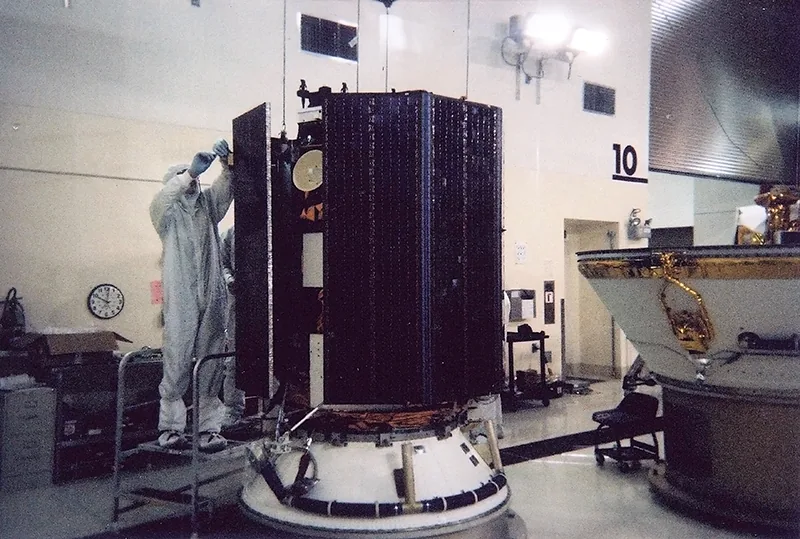 SAC-C stands on a white adapter in a cleanroom, with dark folded solar panels and a technician on a platform alongside it.