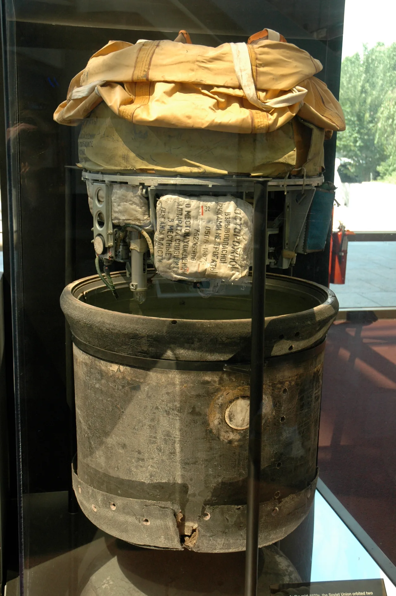 A cylindrical return capsule is displayed opened vertically, exposing its metal framework, fabric packages and a worn outer shell behind glass.
