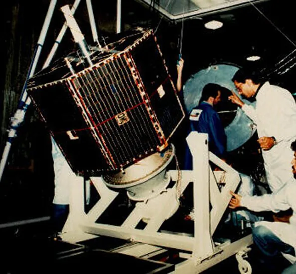 Engineers around a tilted octagonal SCD-1 spacecraft with dark solar-cell panels on a white ground fixture