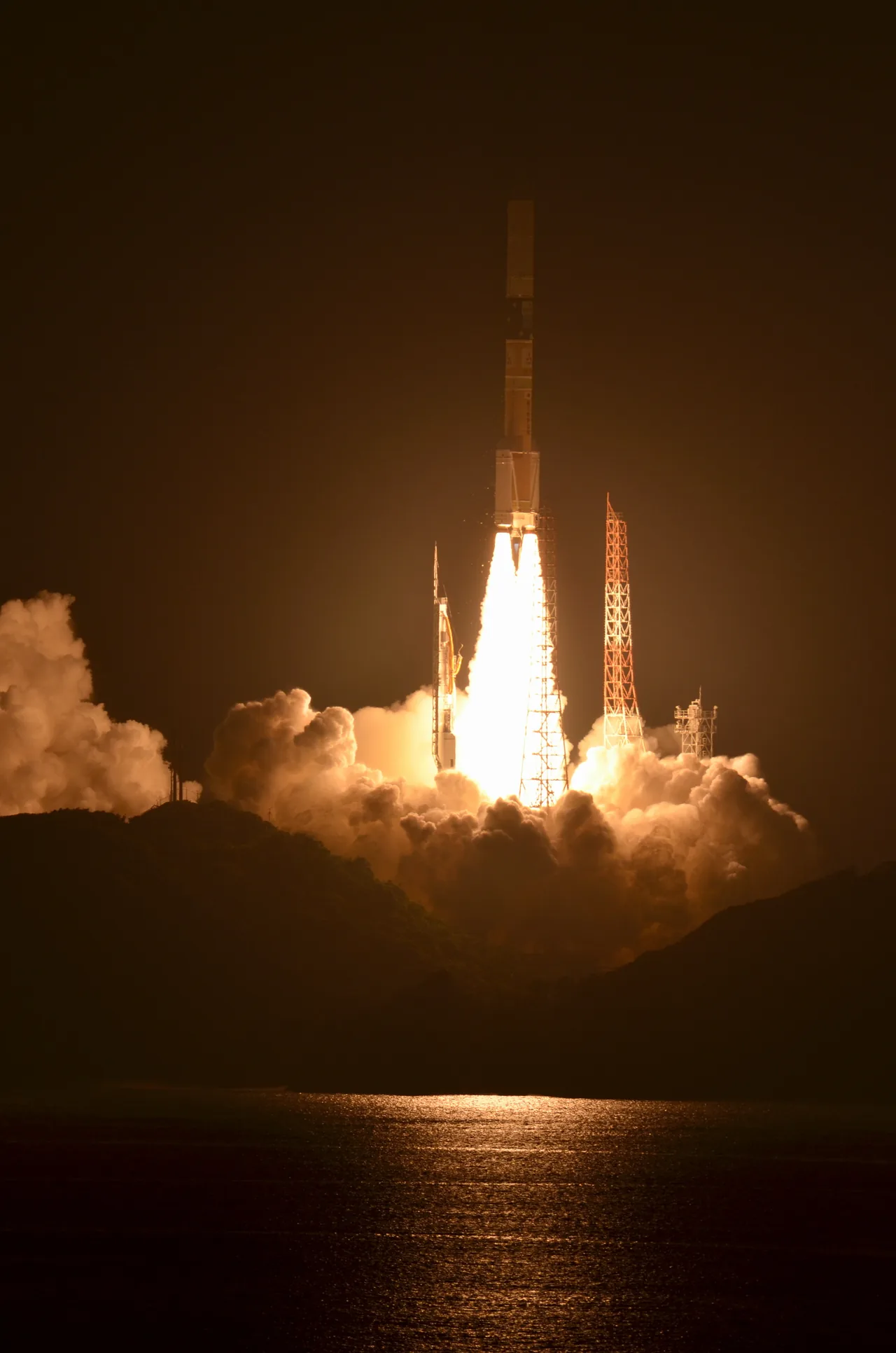 A rocket rises between illuminated launch towers at night, with bright exhaust and smoke reflecting in the water below a dark shoreline.