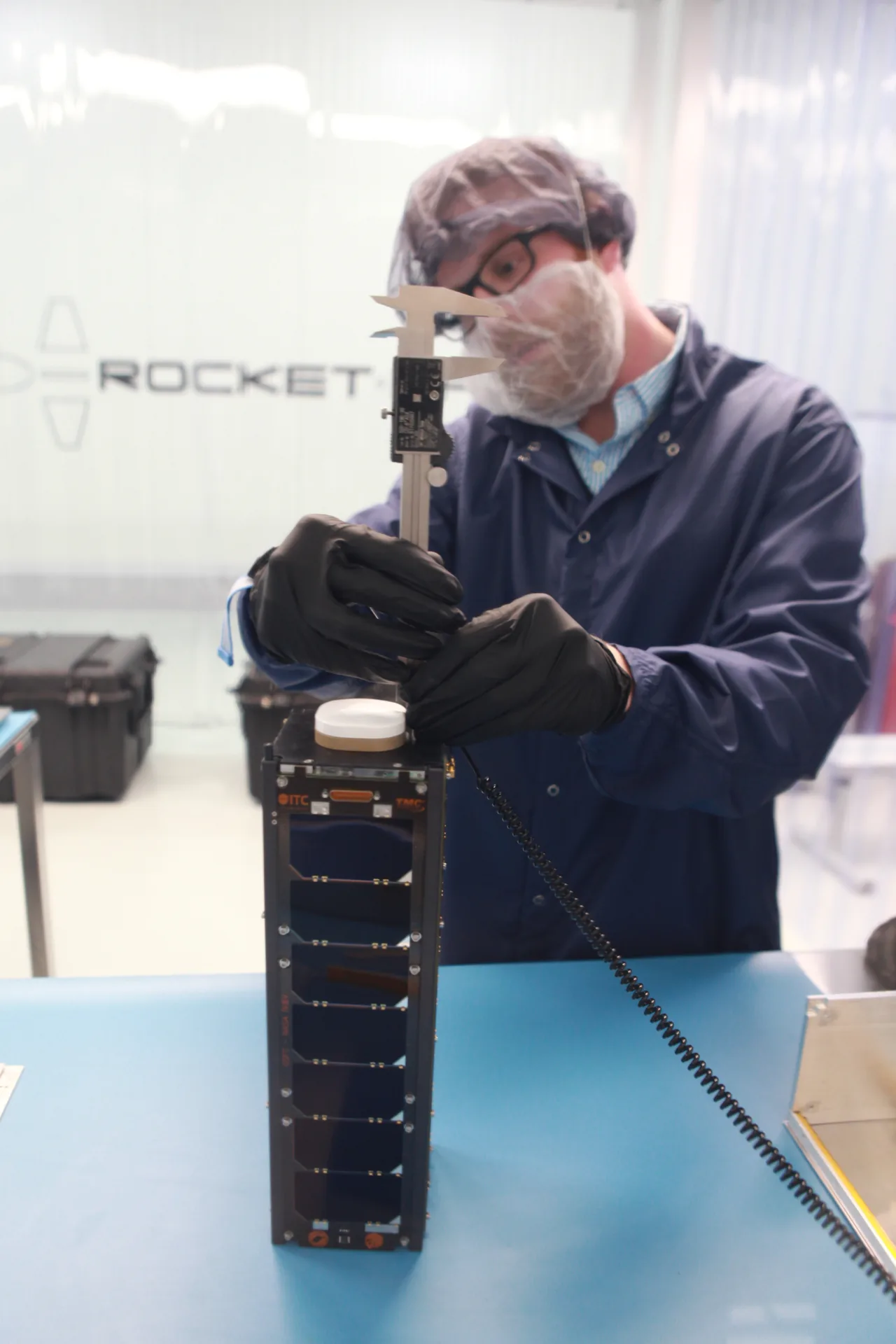 A gloved technician uses calipers to measure a tall, dark CubeSat standing on a blue work surface