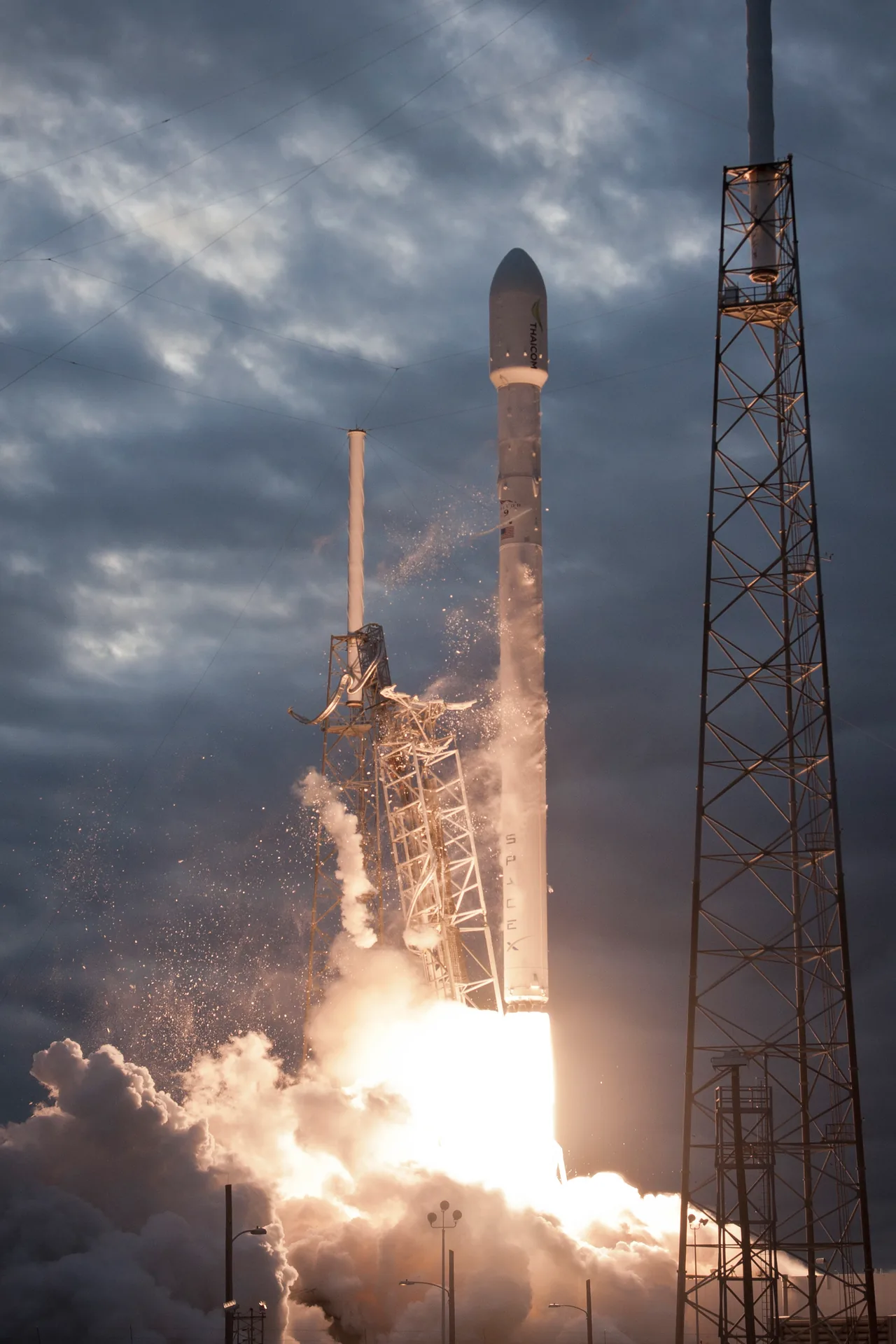 A Falcon 9 lifts beside its launch tower with bright exhaust beneath a cloudy sky