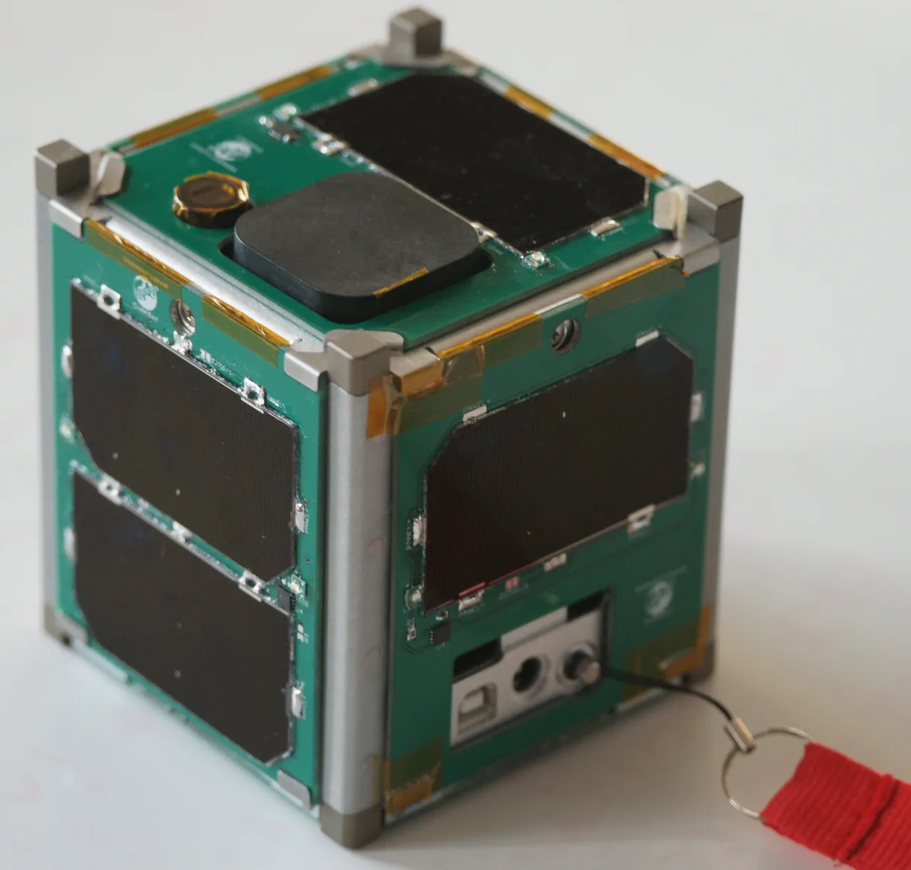 A green CubeSat with dark solar cells and silver corner rails rests on a pale surface, with a red safety-pin tag extending to one side