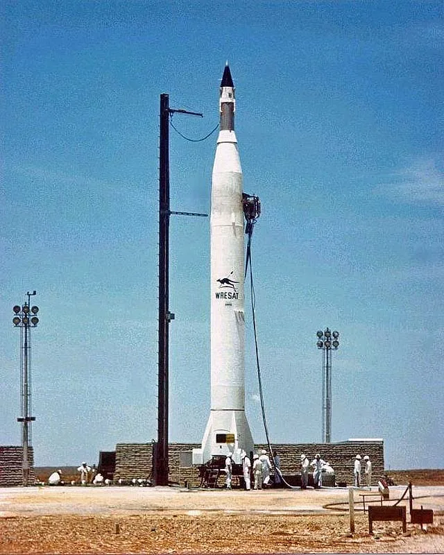 White Redstone-Sparta rocket marked WRESAT beside a slender service mast, with ground personnel at its base.