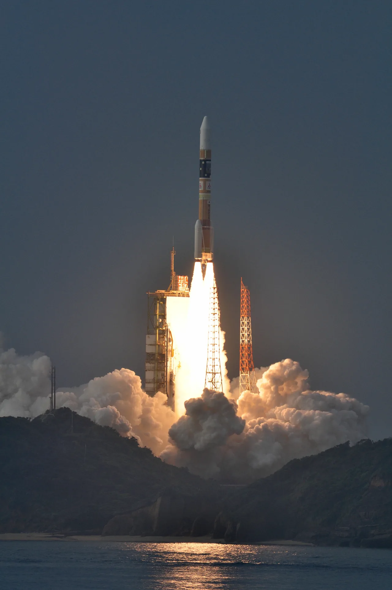 A white and orange rocket rises above a coastal launch pad amid bright exhaust and a broad cloud of smoke