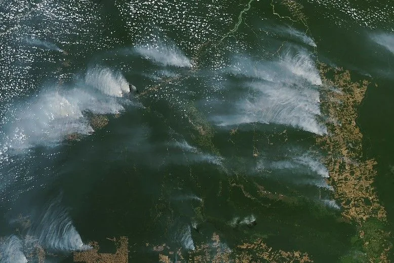Pale smoke plumes spread across dark green forest and tan cleared strips, with scattered white clouds over the Brazilian Amazon