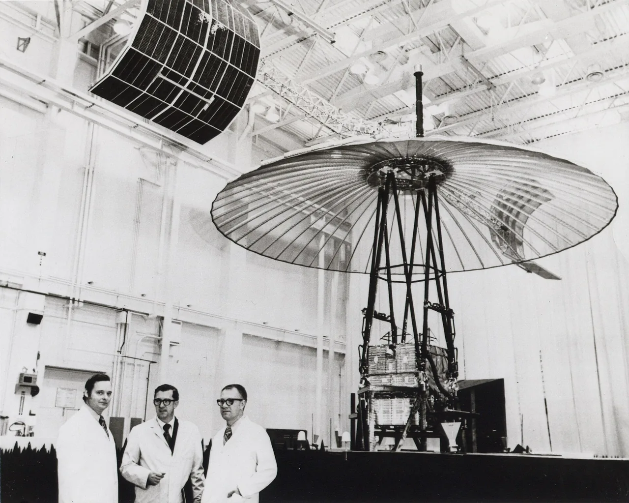 Three people in white coats stand beside ATS-6 and its large dish-shaped reflector in a cleanroom