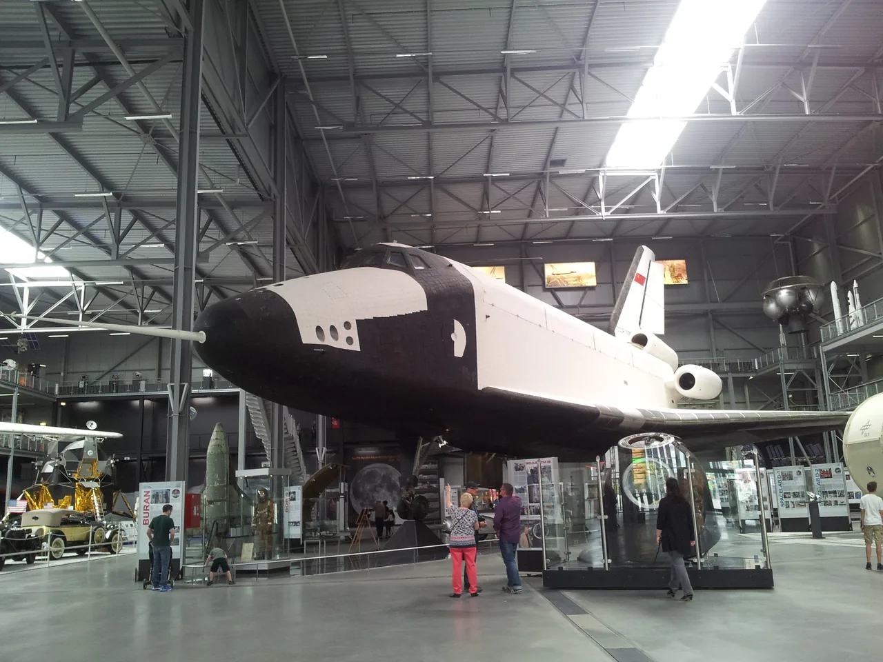 The black-and-white OK-GLI spaceplane displayed inside a museum hall with visitors beneath its nose