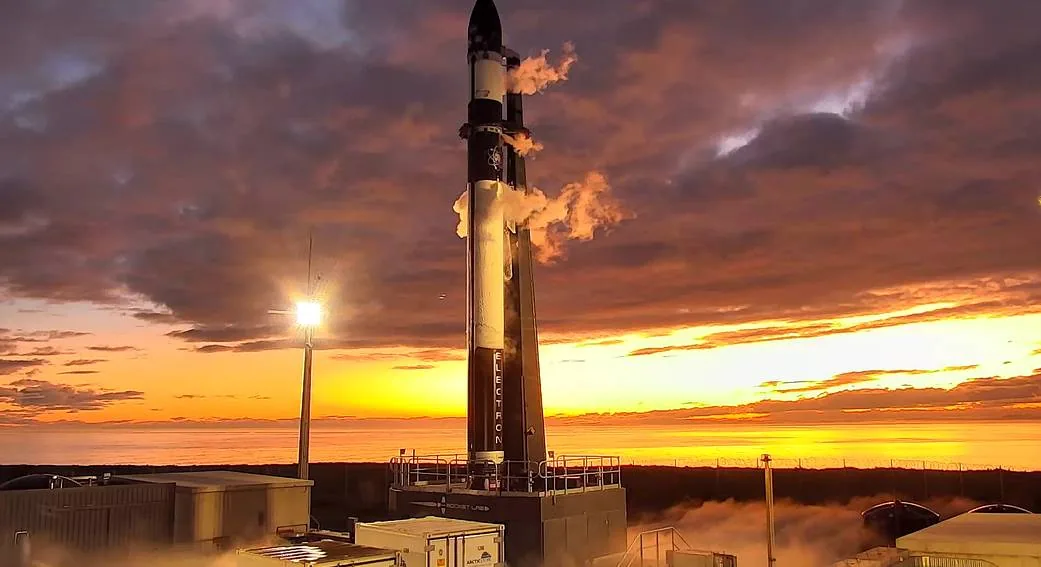 Electron stands on its Māhia launch pad beneath an orange sky, with ground support structures and the enclosed CAPSTONE payload